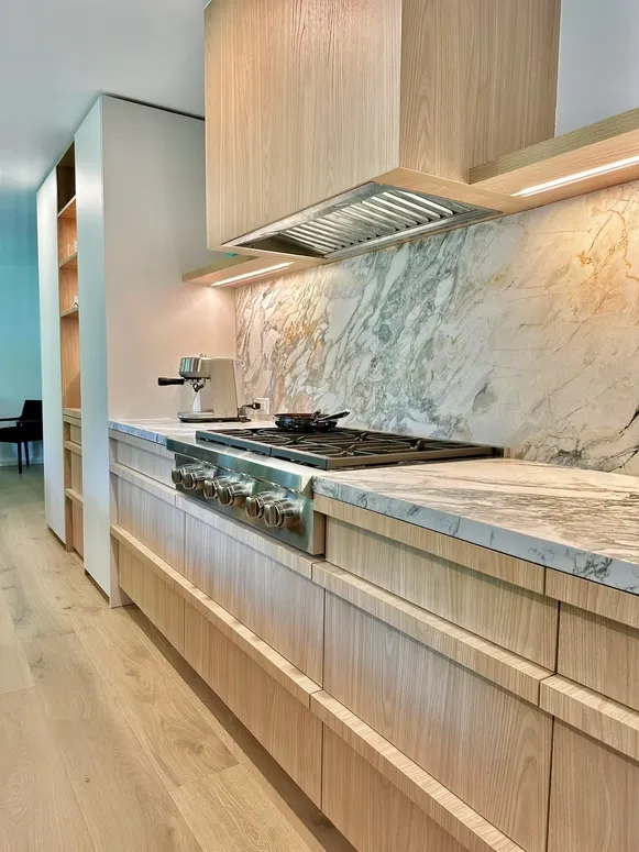 Light oak kitchen with a stone slab backsplash, gas cooktop and an oak range hood with under-cabinet lighting