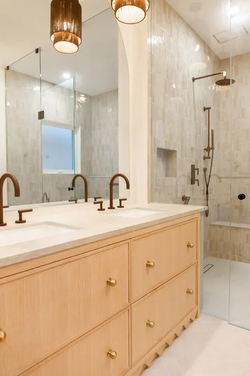 Double-sink light oak vanity with brass knobs beside a glass shower with beige tile