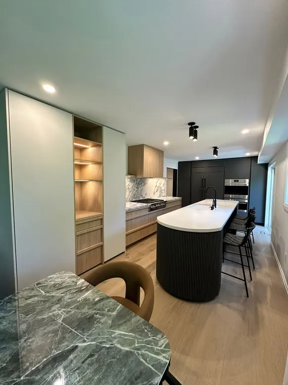Long kitchen with lit oak display shelves, a curved black fluted island and a green marble table