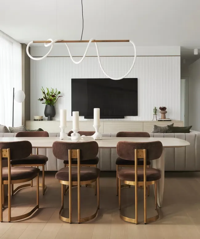 Dining area with an oval table and brown velvet chairs in front of a fluted wall with a TV and credenza