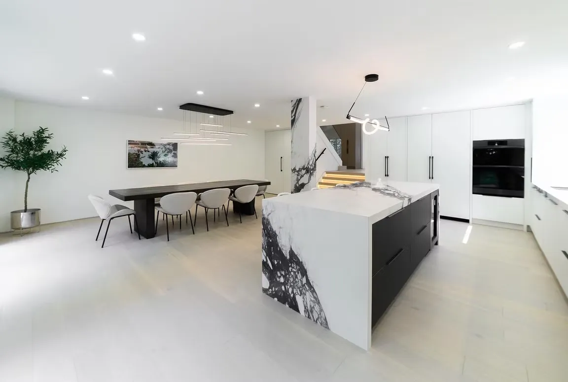 Large white kitchen with a marble waterfall island, a long dining table and built-in ovens